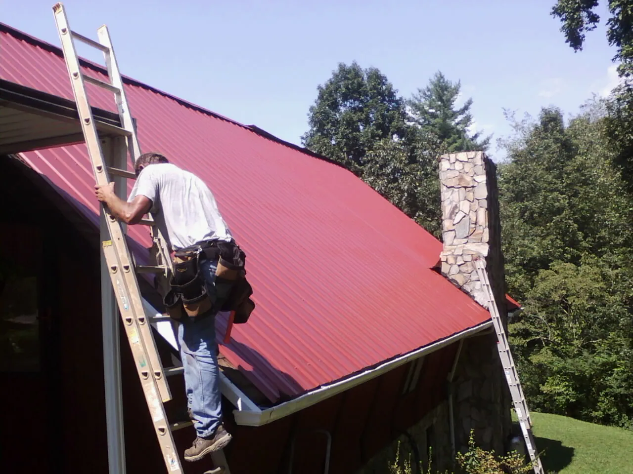 Roofer on a ladder beside a red metal roof and stone chimney — Soffit & Fascia Repair by Roofing Brothers in Granville